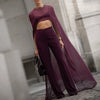 A model wears a sheer, long-sleeved plum mesh poncho cape over a fitted purple jumpsuit in an elegant setting.