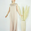 A woman models a flowing, light beige modest abaya dress against a white background with tall ornamental grasses.