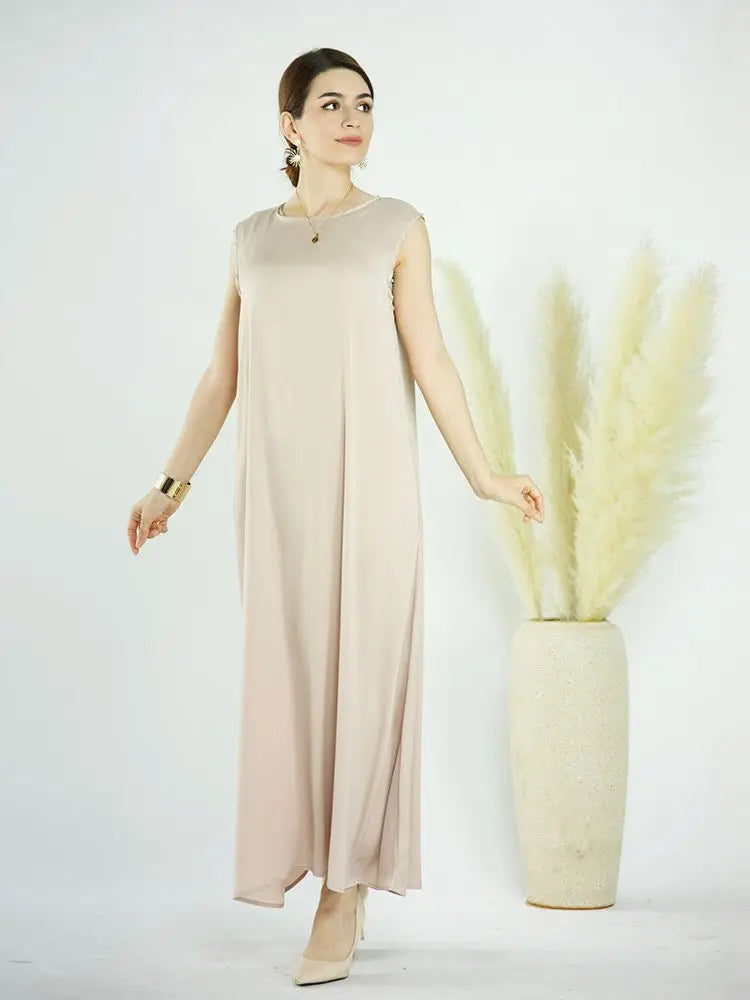 A woman models a flowing, light beige modest abaya dress against a white background with tall ornamental grasses.