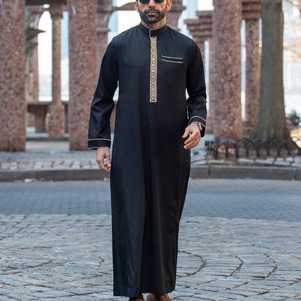 Mann in schwarzem besticktem Langarm-Arabischer Thobe im Freien vor architektonischer Kulisse.