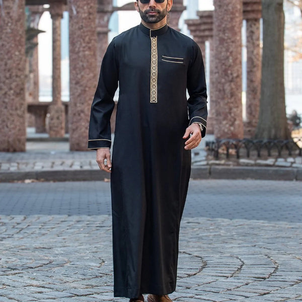 A man wearing a black embroidered long sleeve Arabian thobe standing outdoors in an architectural setting.
