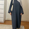 Une femme portant une abaya ample de couleur unie élégante et un hijab gris clair se tient dans une pièce bien éclairée.