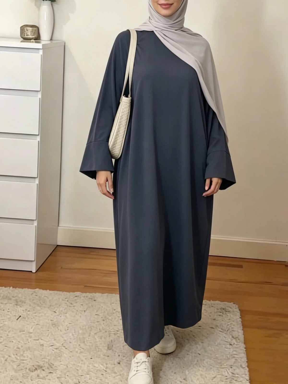 Une femme portant une abaya ample de couleur unie élégante et un hijab gris clair se tient dans une pièce bien éclairée.