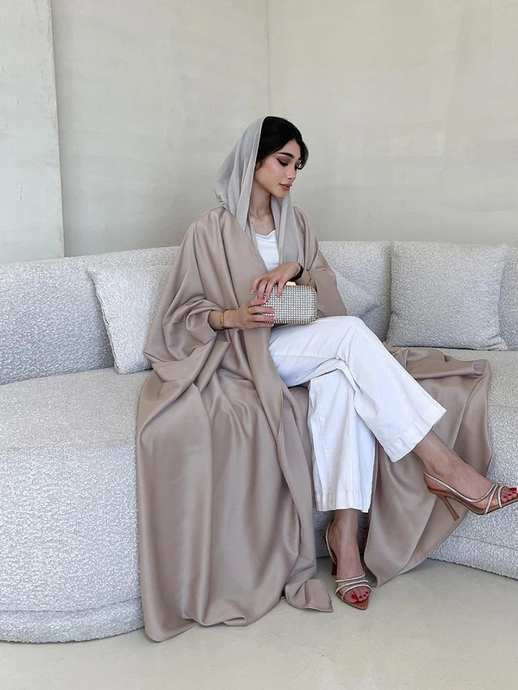Una mujer vistiendo una abaya de satén beige con mangas de murciélago y pantalones blancos, sentada en un sofá.