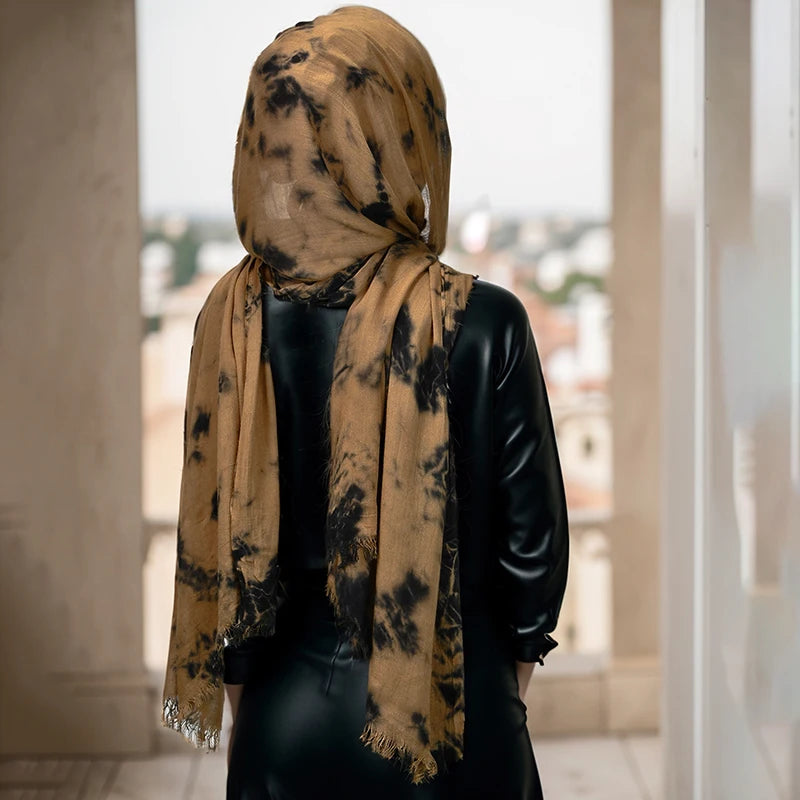 Eine Frau mit gelb-braunem Tie-Dye-Hijab-Schal über einem schwarzen Oberteil, steht drinnen am Fenster.