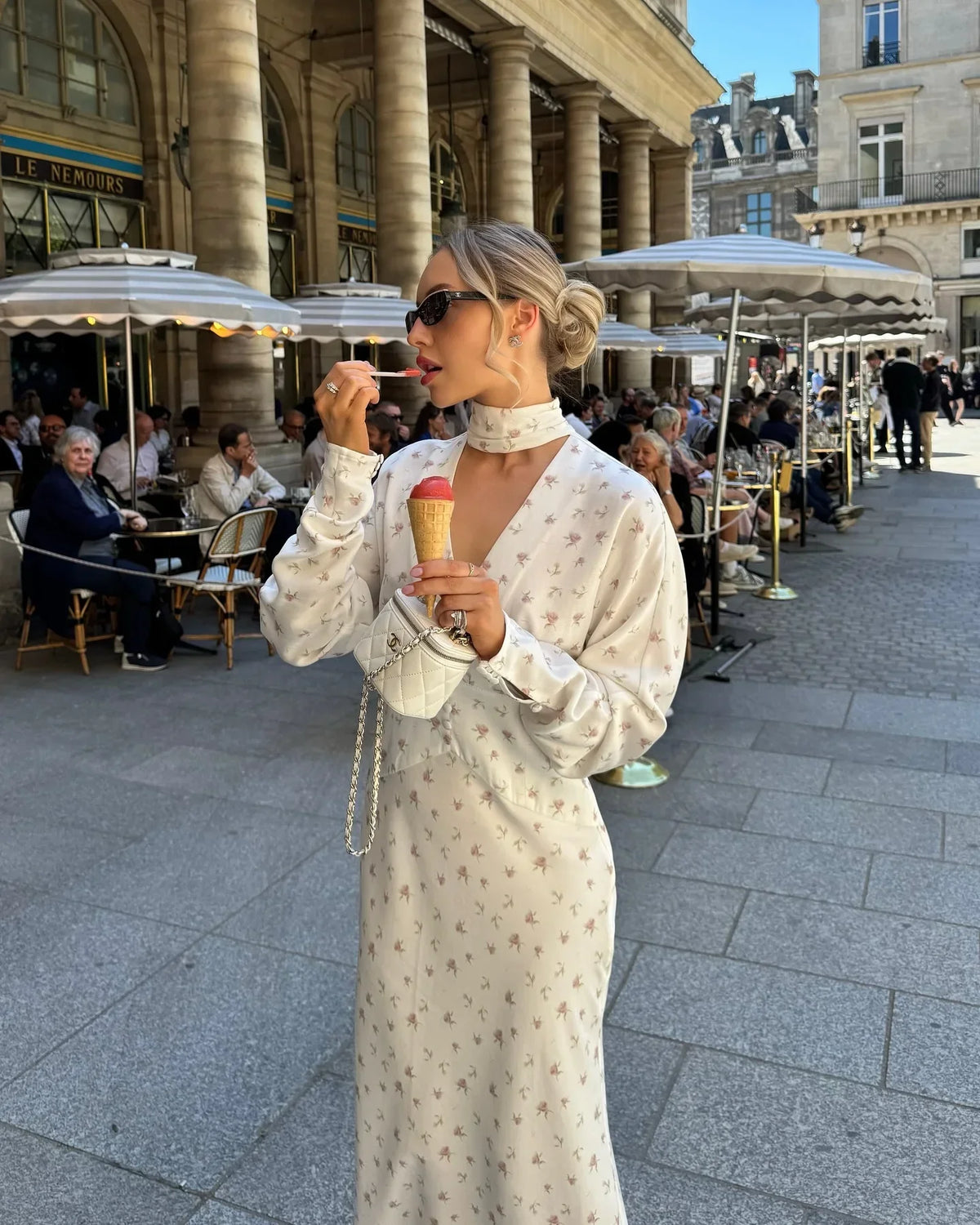 Una mujer vistiendo un vestido largo de satén con estampado floral y gafas de sol está al aire libre en una plaza pública.