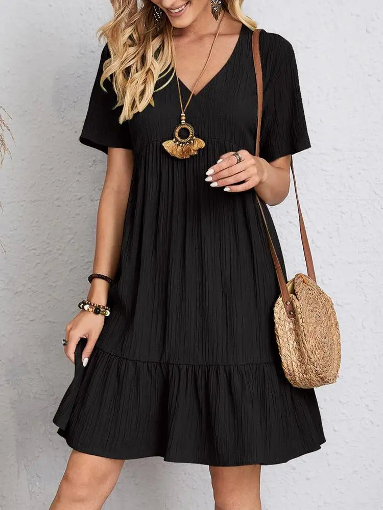 Eine Frau modelliert ein elegantes, schwarzes, A-linienförmiges Mini-Kleid mit goldener Halskette und Stroh-Schultertasche.