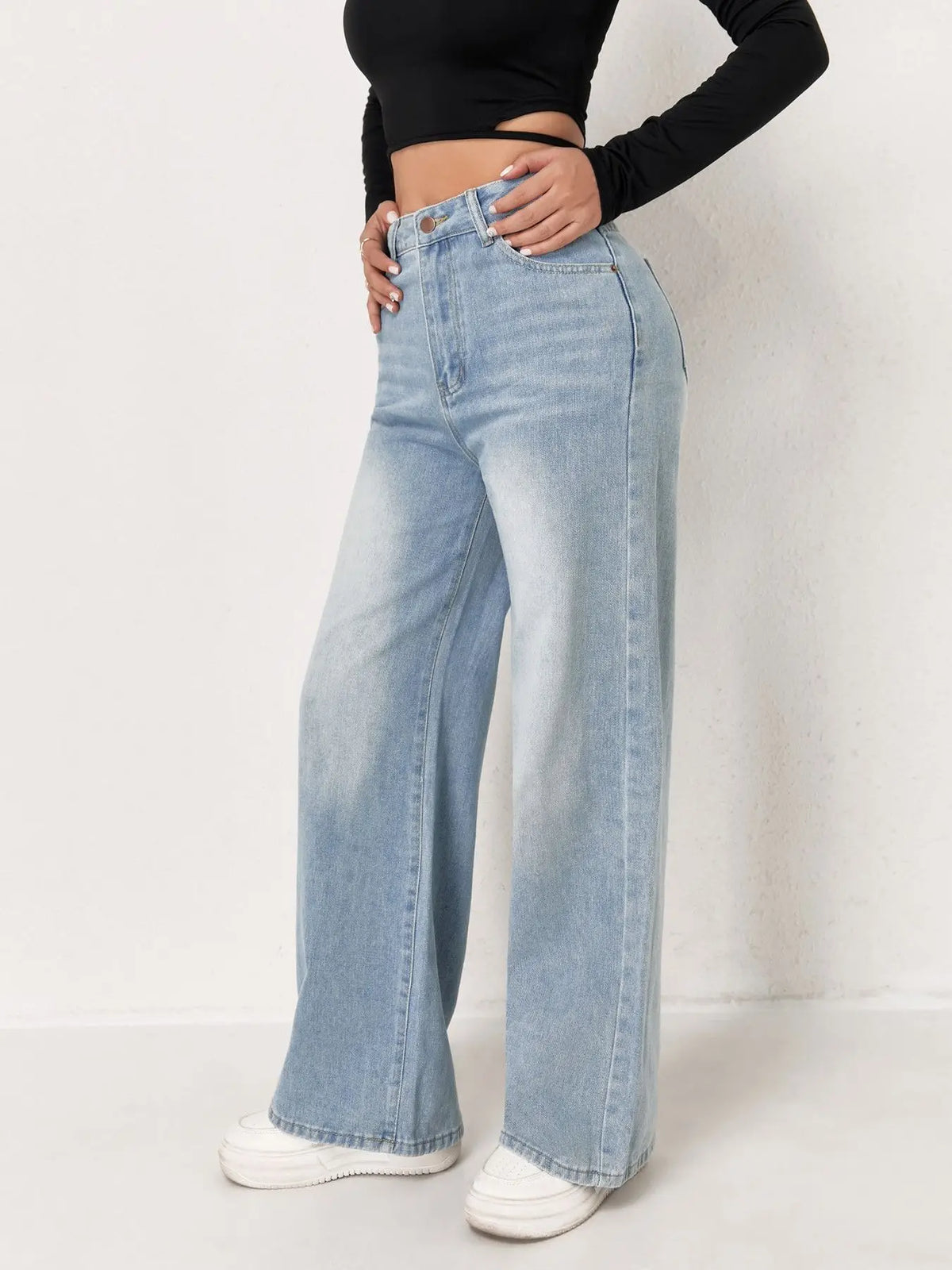 Una mujer vistiendo pantalones vaqueros anchos de color azul claro y una blusa negra, de pie en interiores.