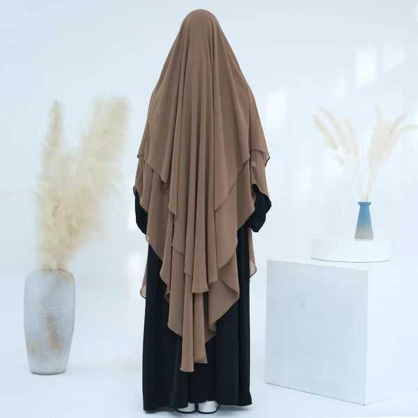 A brown two-layer chiffon niqab headscarf displayed against a white background with dried floral arrangements.