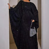 Modelo vistiendo una abaya negra de manga de murciélago de lujo con brillo sutil, de pie en un ambiente interior luminoso.