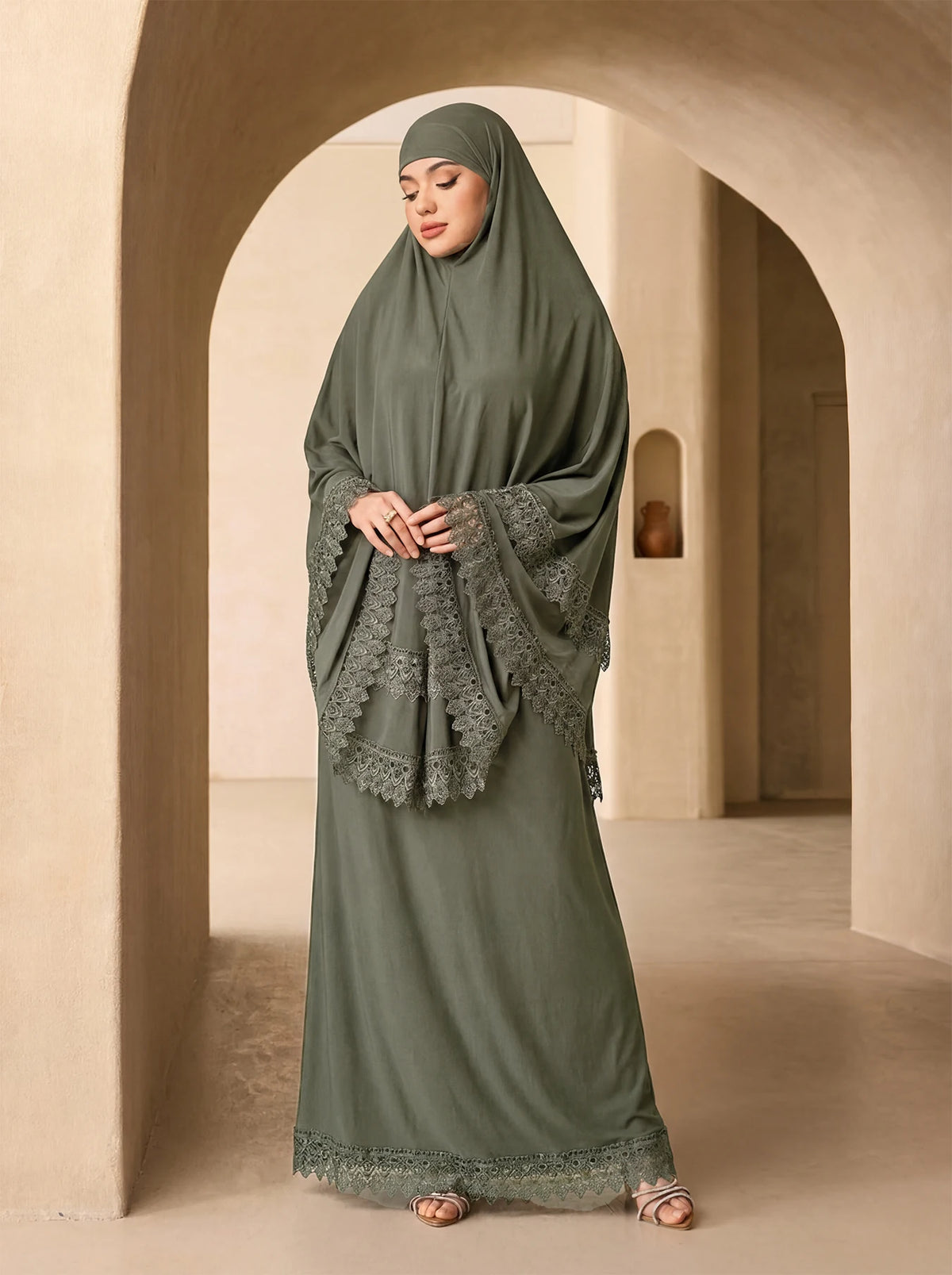 Eine Frau in einem eleganten, mehrlagigen Spitzen-Khimar-Abaya-Set in Olivgrün steht in einem hellen Gewölbe-Flur.