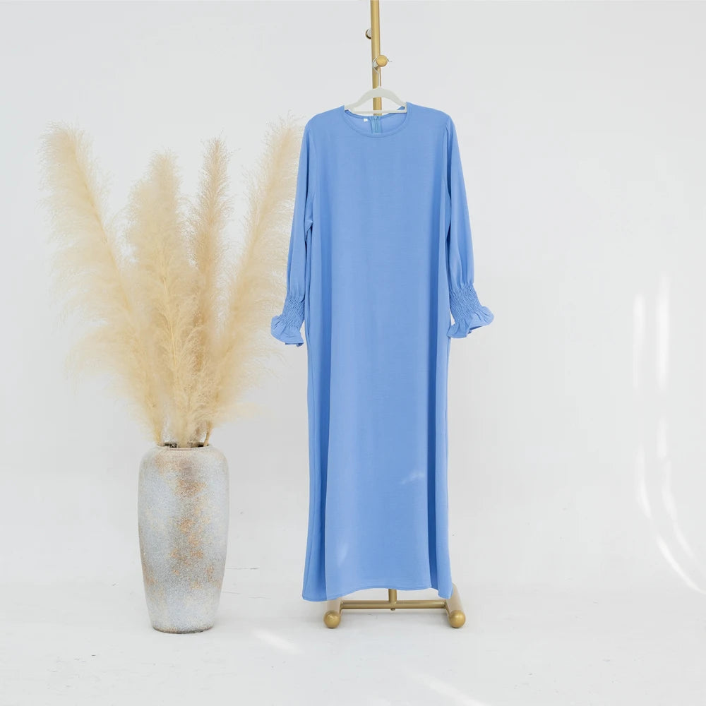 Vestido abaya azul claro y holgado colgado en una percha junto a un jarrón de pampas grass beige seco.