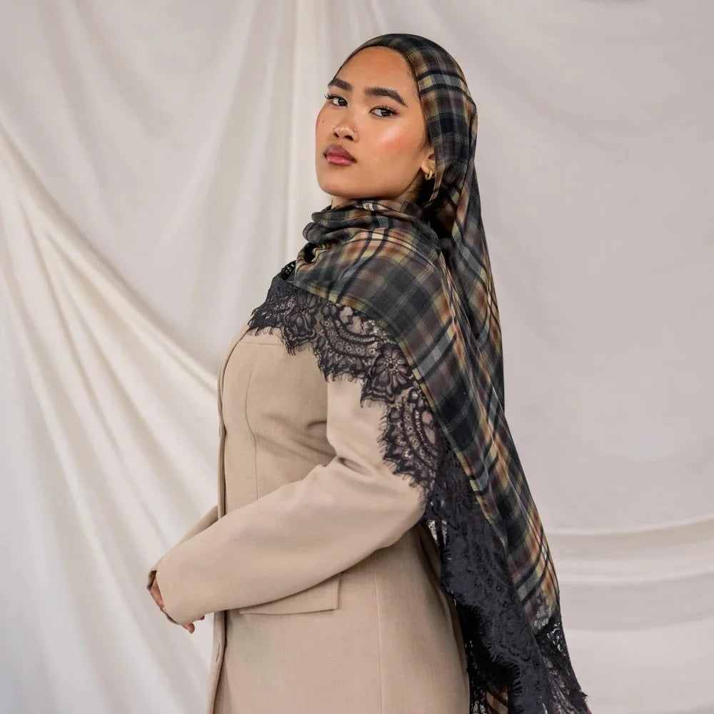 A woman wearing a beige blazer and a plaid hijab scarf with black lace detailing against a white background.