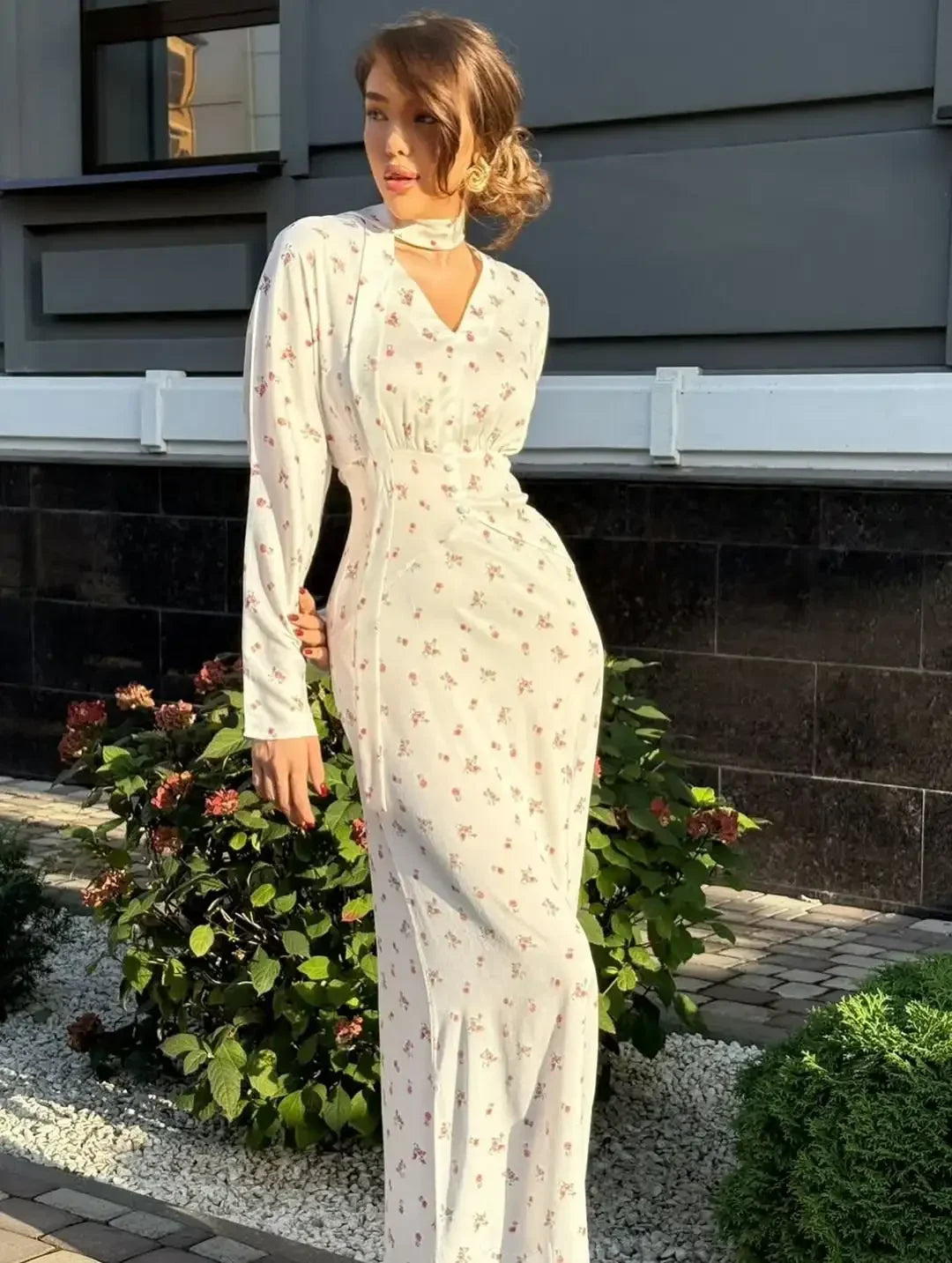 Una mujer vistiendo un vestido largo de satén blanco con estampado floral, de manga larga, de pie al aire libre contra una pa
