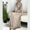 Una mujer vistiendo un abaya maxi de satén beige fluido y un hiyab a juego, sentada en interiores.