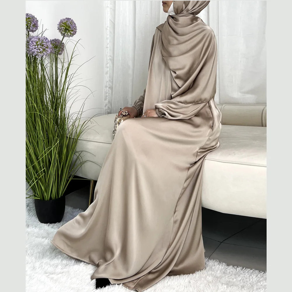 Una mujer vistiendo un abaya largo de satén beige y hijab a juego, sentada en interiores.