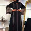Una mujer vistiendo un vestido abaya negro de manga larga y puños bordados, de pie frente a un espejo con marco de oro.