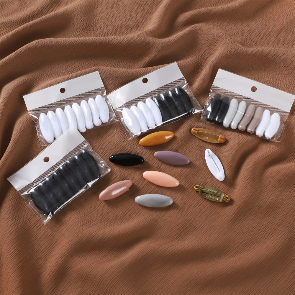A collection of eight solid color brooch pins in various shades of black, white, and nude are displayed in clear packaging on