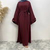 Una elegante abaya de encaje color granate para mujeres, exhibida contra una pared blanca sobre un suelo gris texturizado.
