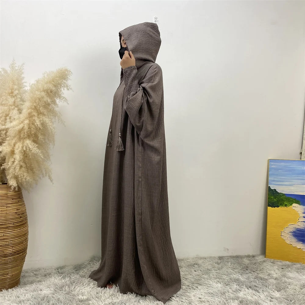 Une longue abaya gris foncé avec capuche et manches batwing, exposée contre un mur blanc.