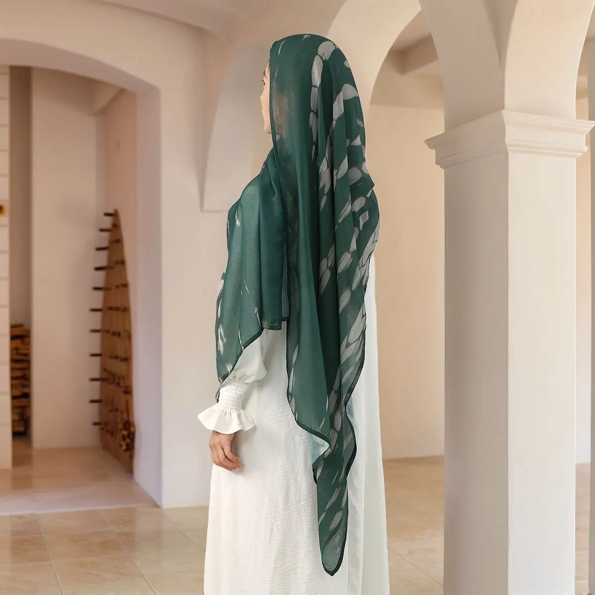 Eine Frau in einem eleganten grünen und weißen Kleid mit Hijab steht in einem hellen, gewölbten Flur.
