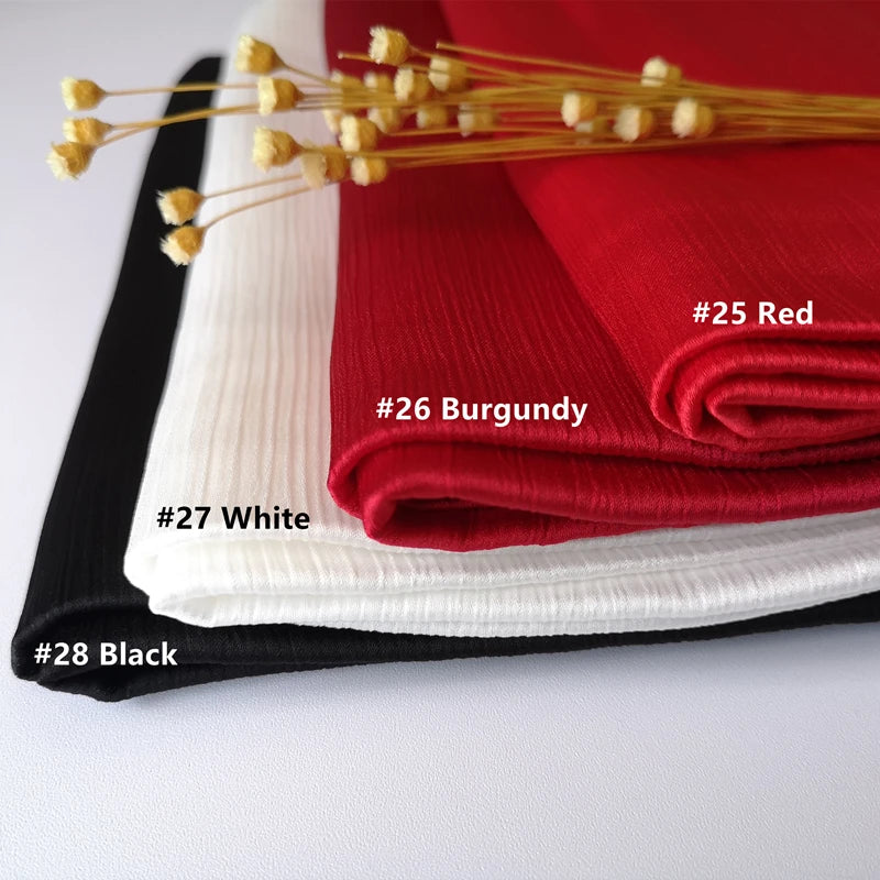 Ein Stapel aus gerüschtem Satin-Chiffon-Hijabs in Rot, Bordeaux, Weiß und Schwarz auf einer weißen Oberfläche.