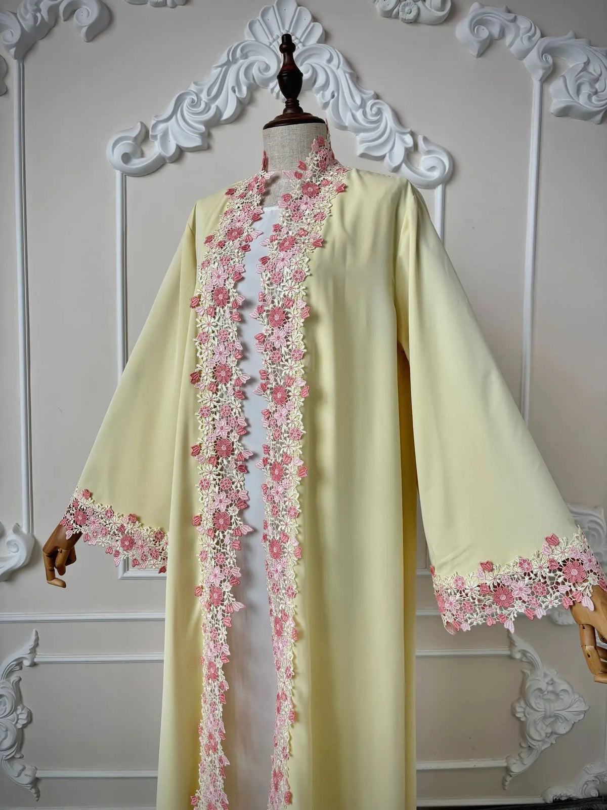 Une longue abaya jaune pâle avec des bordures de dentelle florale rose délicates sur le col et les poignets.