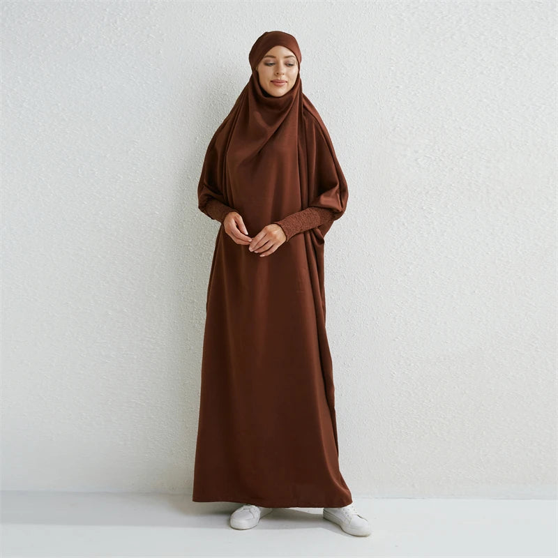 Eine Frau in einem braunen, bescheidenen Abaya mit gerafften Ärmeln vor hellem Hintergrund.