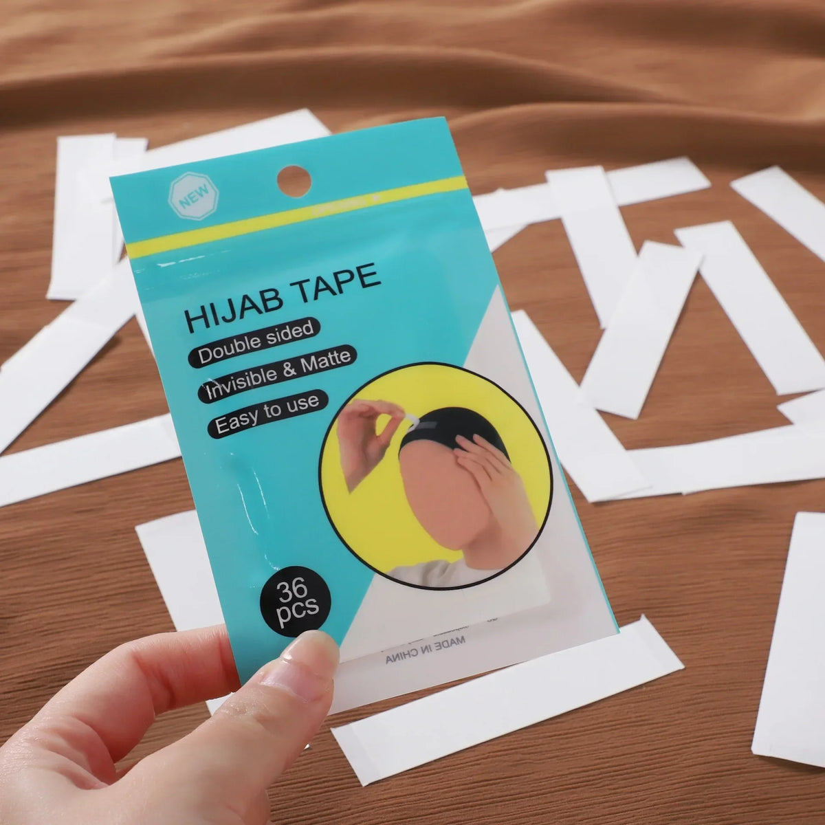 Una mano sostiene un paquete de cinta para hijab invisible mate de doble cara con 36 piezas sobre una superficie marrón.