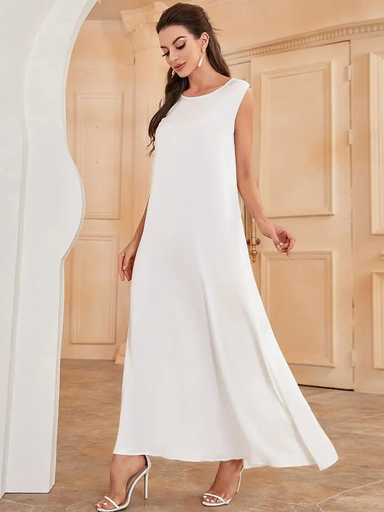 A woman models a white flowing modest abaya dress with long sleeves and a high slit in an indoor setting.