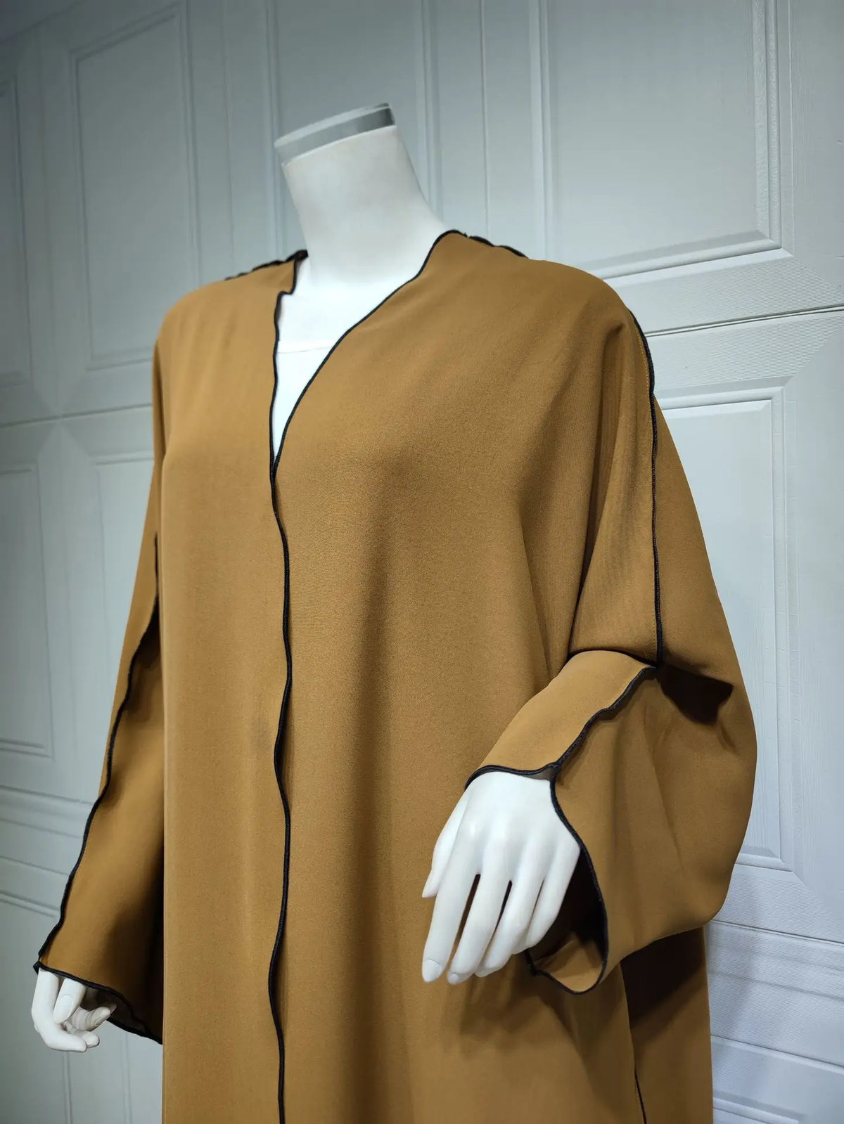 Una modelo viste un amplio vestido abaya marrón hasta el suelo con ribetes negros contra un fondo blanco acanalado.