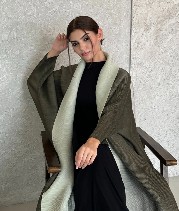 A woman wearing a long, olive green pleated abaya cardigan over a black dress, standing against a light textured wall.