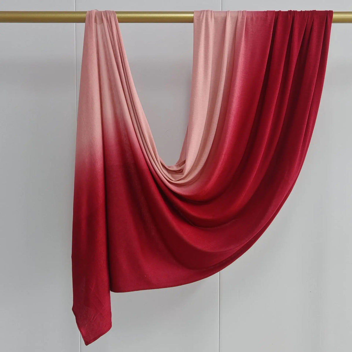 Pañuelo hijab de algodón degradado doble cara en tonos rosa y rojo, drapeado sobre una varilla dorada.