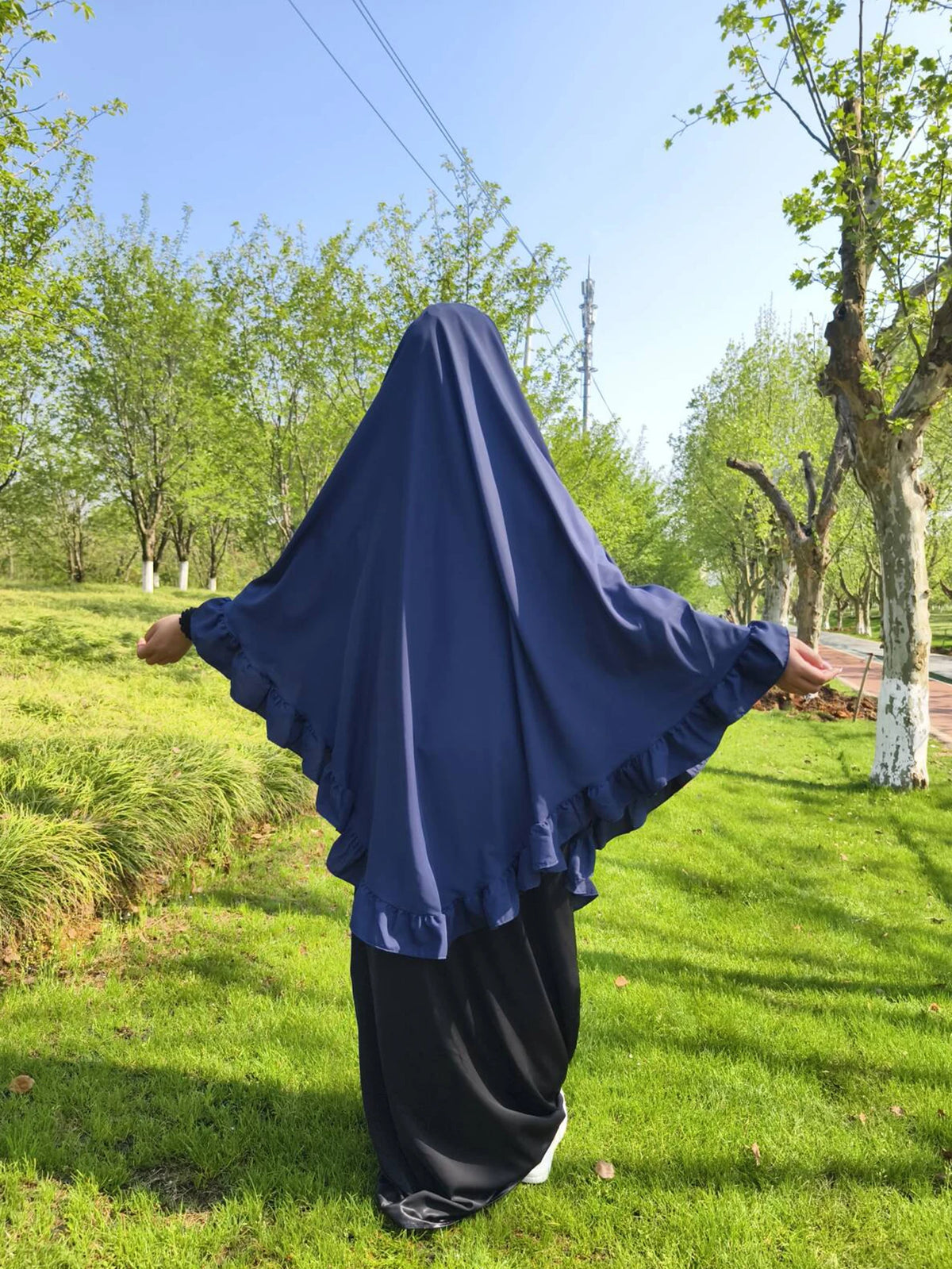 Eine Frau mit dunkelblauem Diamant-Hijab steht draußen in einem grasbewachsenen Park.