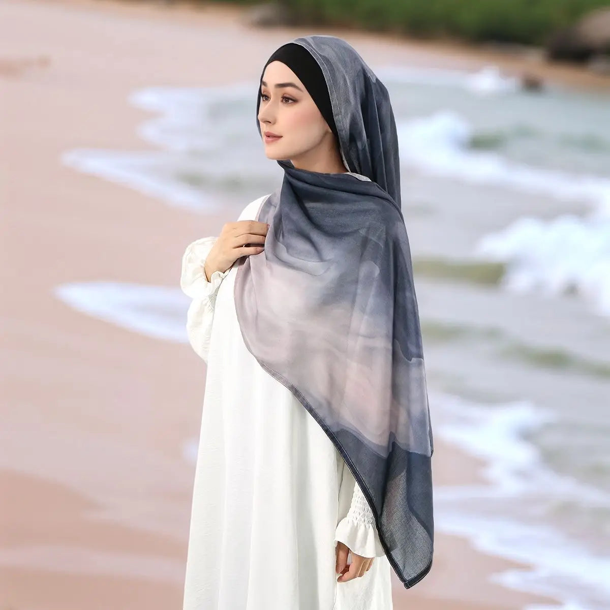 Eine Frau trägt einen durchscheinenden grauen Aquarell-Digitaldruck-Hijab und weiße, bescheidene Kleidung am Ozean.