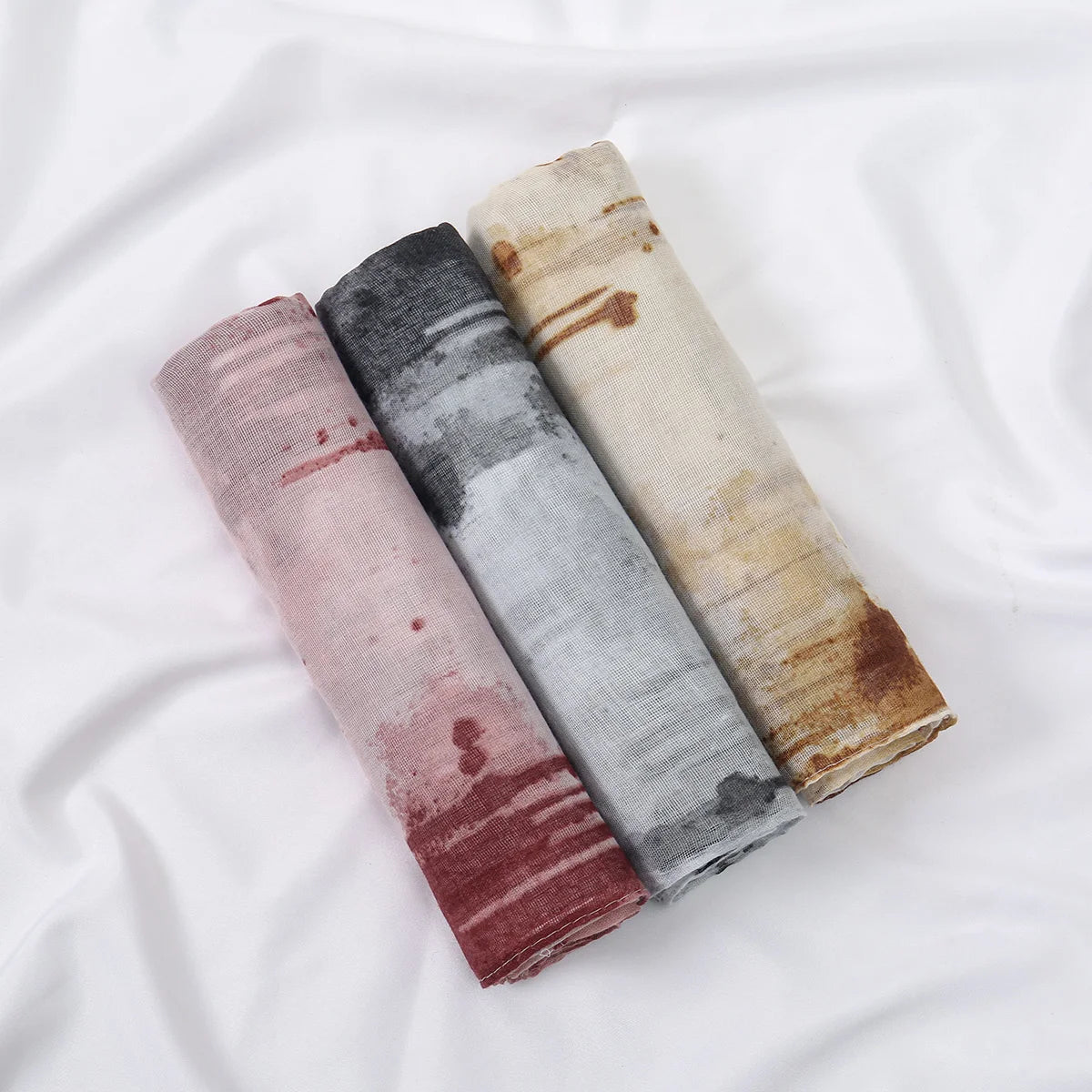 Drei gefaltete, florale Druck-Voile-Schals in Rosa-, Grau- und Beige-Tönen auf weißem Stoff.