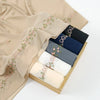 A beige chiffon hijab shawl with delicate pink floral embroidery draped next to a stack of dark blue and navy fabric rolls.