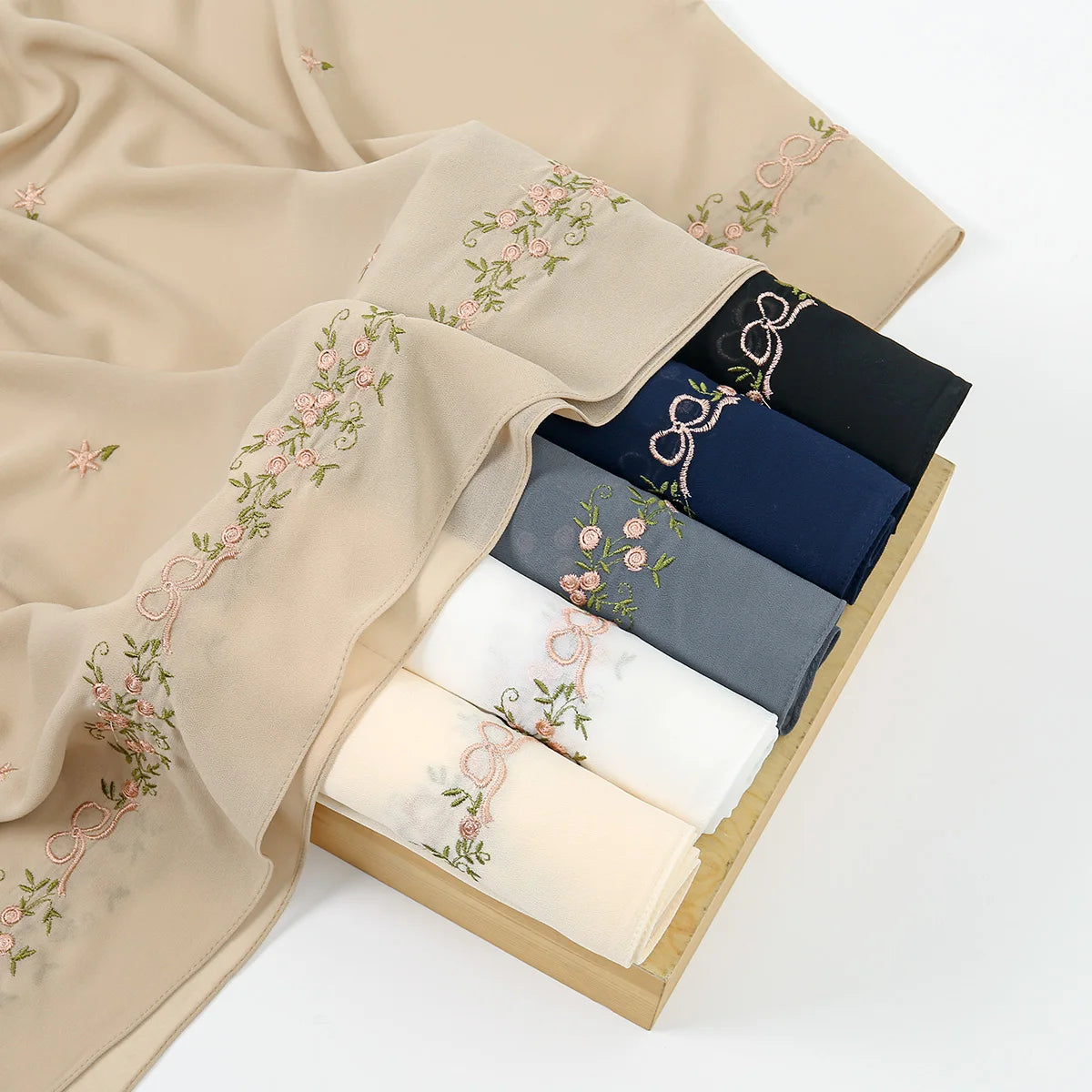 A beige chiffon hijab shawl with delicate pink floral embroidery draped next to a stack of dark blue and navy fabric rolls.