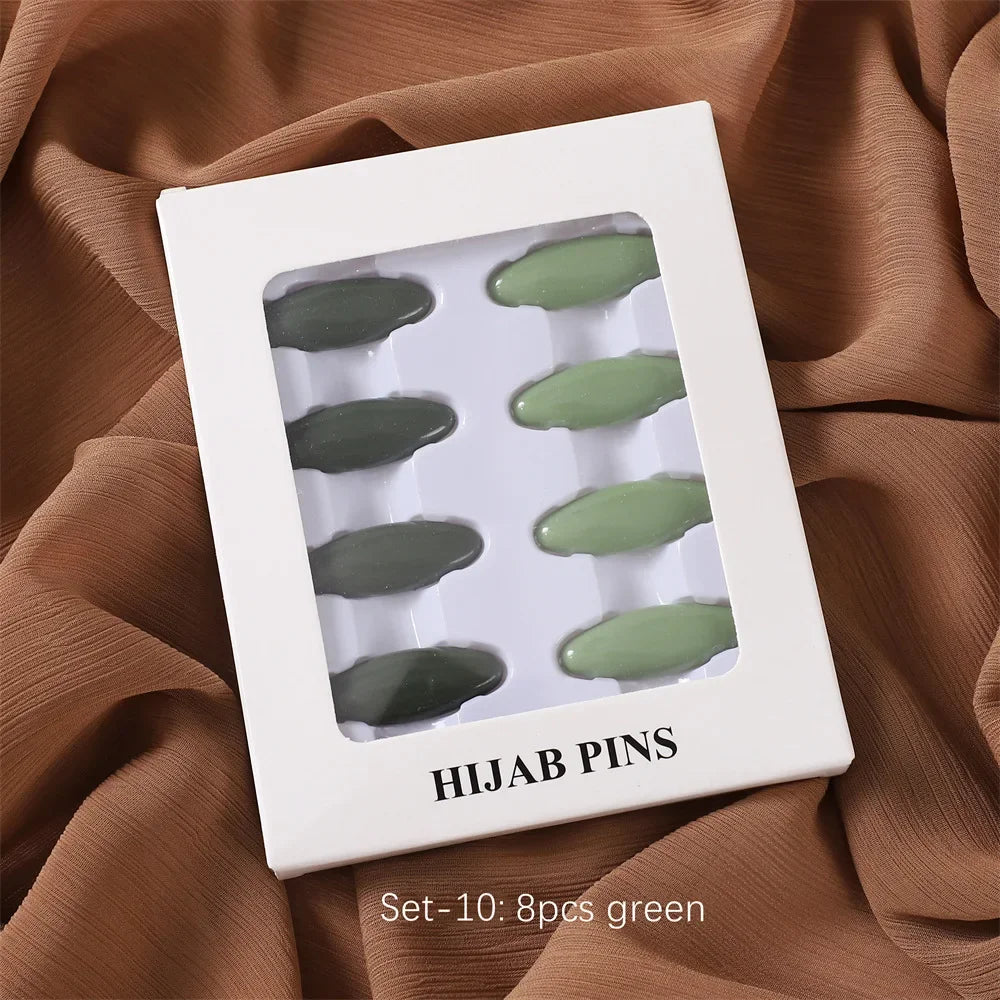 Ein Set von zehn grünen Hijab-Pins in einer weißen rechteckigen Schachtel auf brachem Stoffhintergrund.