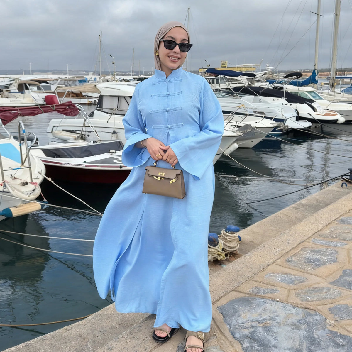 A woman wearing a light blue elegant two-piece Arabic modest set standing on a dock with boats in the background.