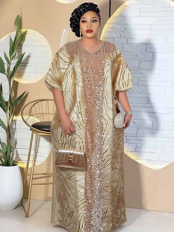 A woman models a gold sequin dashiki maxi dress with a headtie against a white brick background.