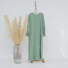 Vestido abaya holgado verde salvia con puños fruncidos colgado en percha de madera.