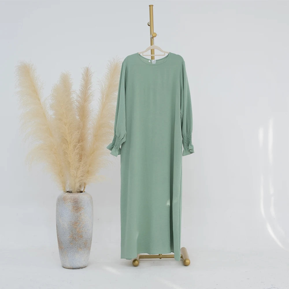 Vestido abaya holgado verde salvia con puños fruncidos colgado en percha de madera.