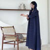 Une femme en manteau long plissé bleu foncé se tient à côté d'une chaise en bois et d'une étagère blanche.