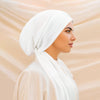 A woman wearing a white solid color turban hijab cap and matching garment, shown in profile.