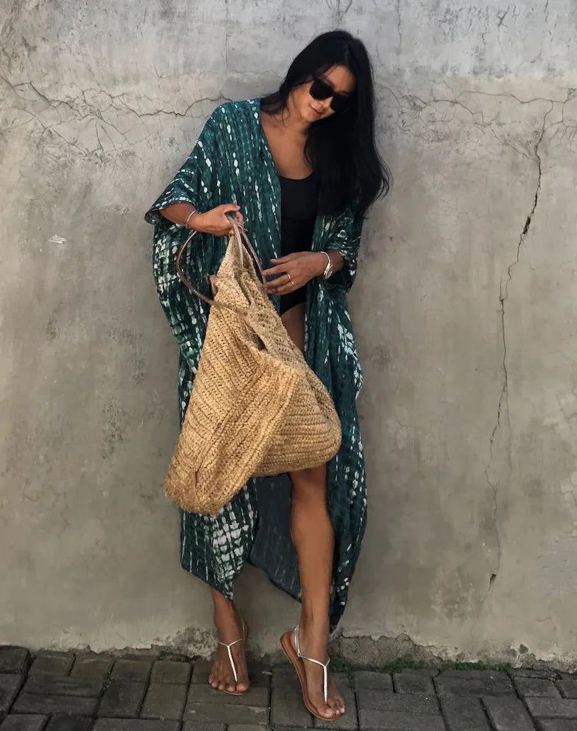 Eine Frau in einem grün gemusterten Kimono-Überkleid und Sonnenbrille steht vor einer Betonwand.