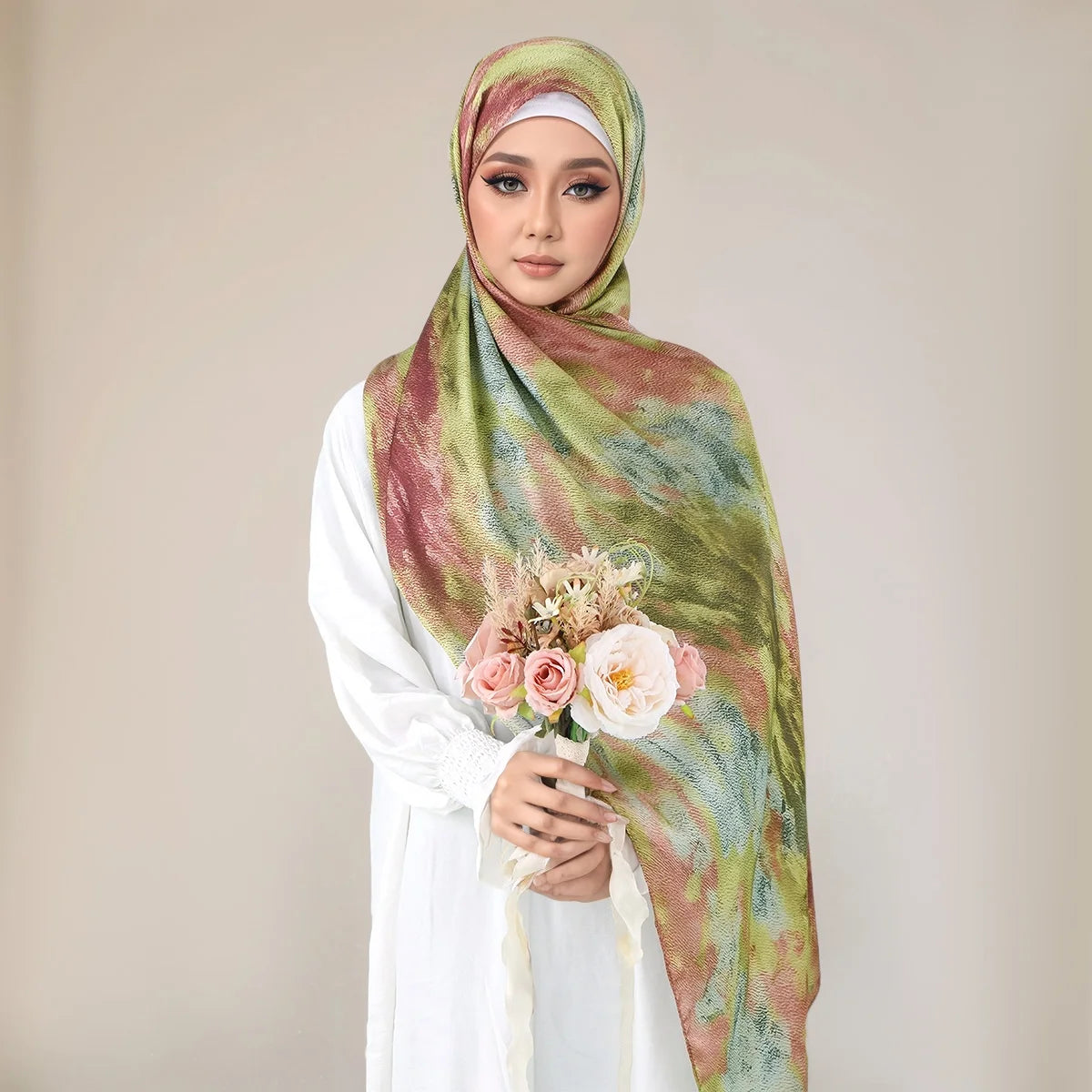 Eine Frau trägt einen eleganten bedruckten Seiderschal-Hijab in Grüntönen, Pink und Gold über einem weißen Outfit.