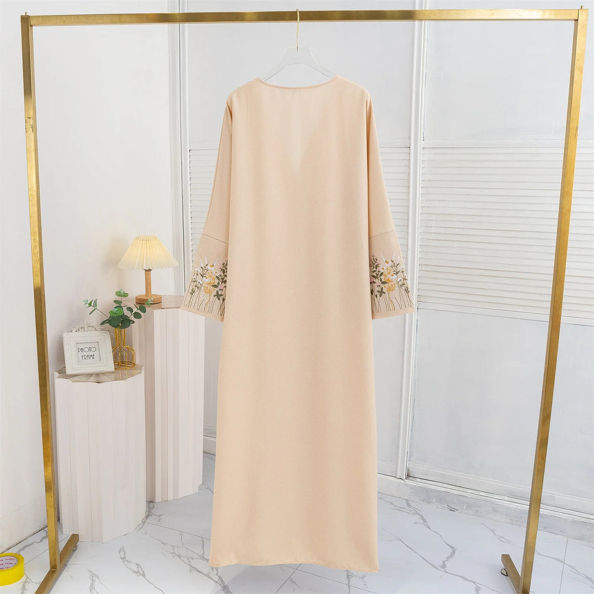 Beige Maxi-Abaya mit Blumenstickerei auf goldener Kleiderstange in einem hellen Raum.