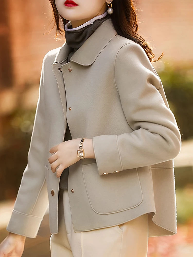 Une femme portant un manteau en laine beige à boutons et un pantalon clair, avec une écharpe grise et un bracelet en or.