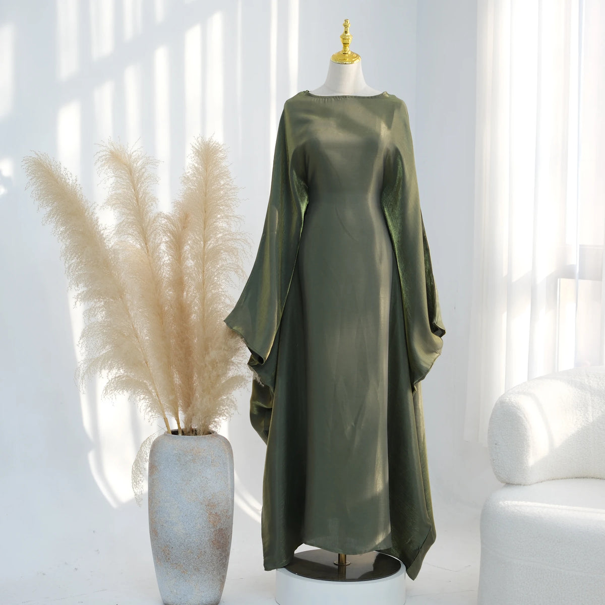 Langes olivgrünes Kaftankleid mit Schmetterlingsärmeln auf einem Mannequin neben getrocknetem Pampasgras.